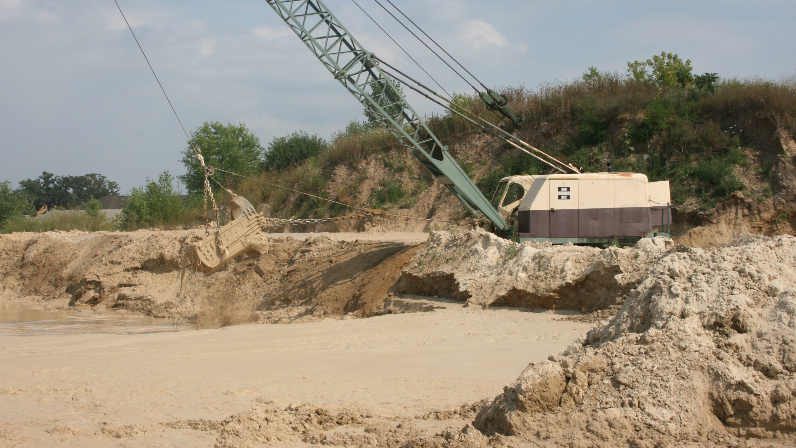 A tan and brown dragline crawler crane dredging a pond with stacked sand in the foreground.