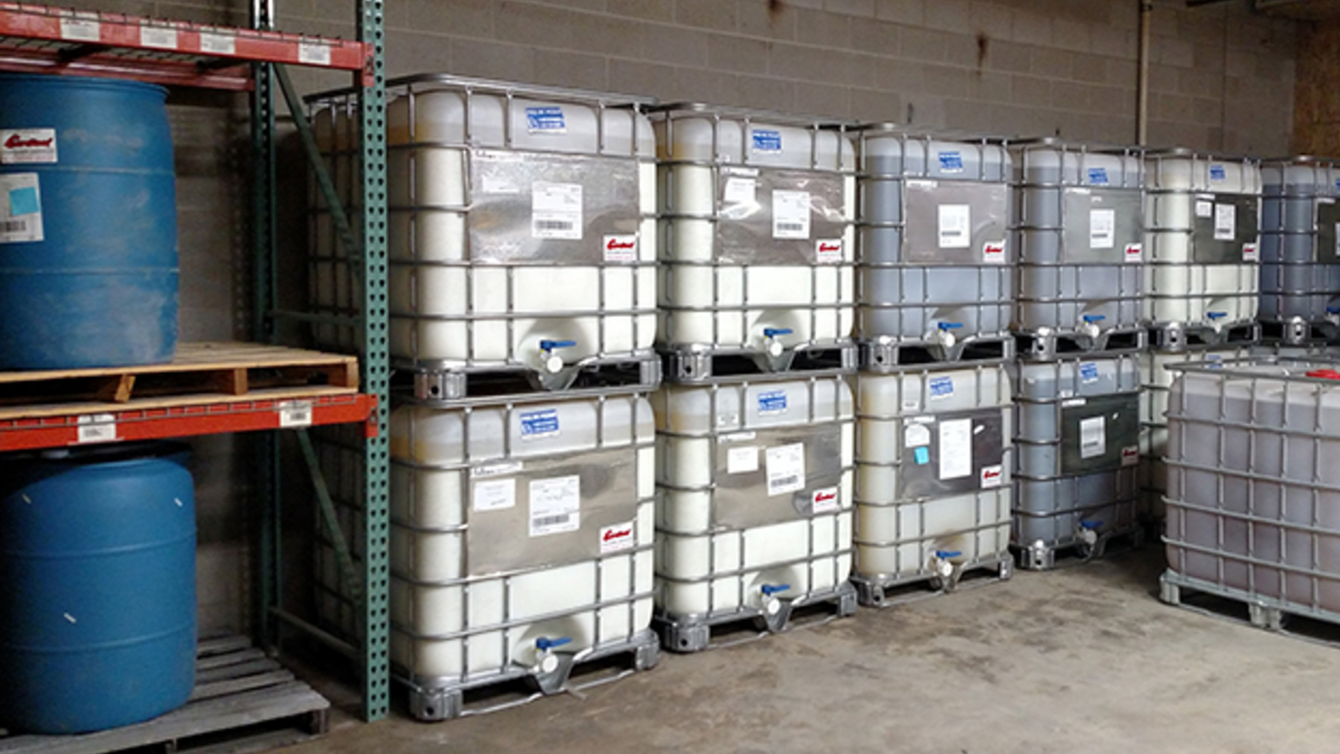 A picture showing liquid polymer totes stacked on top of each other and liquid polymer drums on shelving in a warehouse.