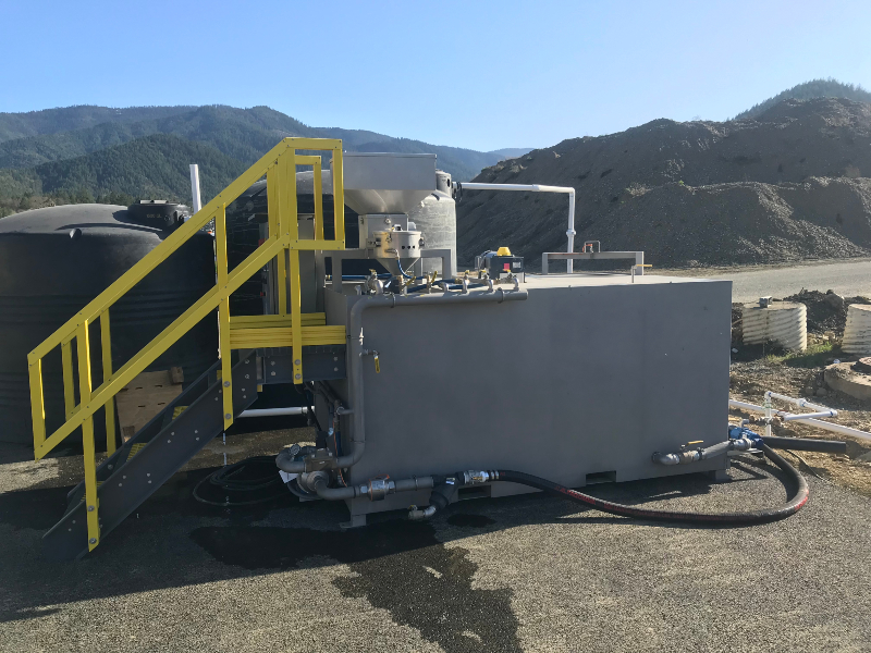 The side of a dry polymer unit at a sand and gravel site with water tanks behind it and mountains in the background.