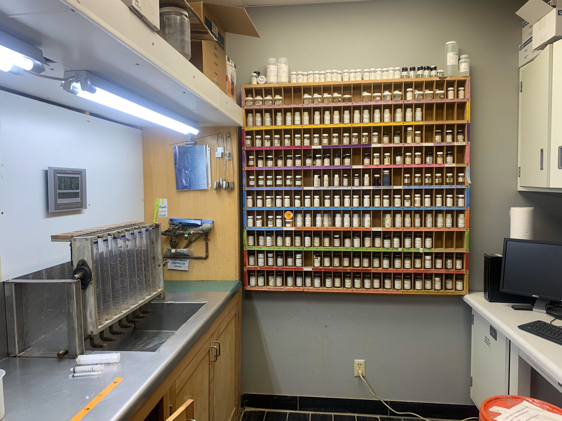Una habitación pequeña con vasos de precipitados sobre el fregadero de la izquierda y una pared de diferentes productos químicos en el fondo.