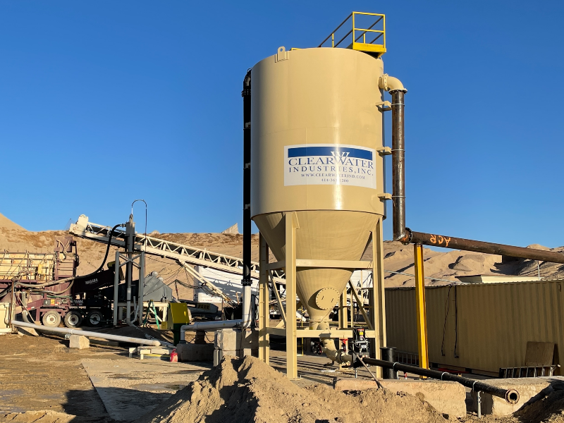 A large, tan, and vertical clarifier tank in the sun with aggregate equipment in the background.