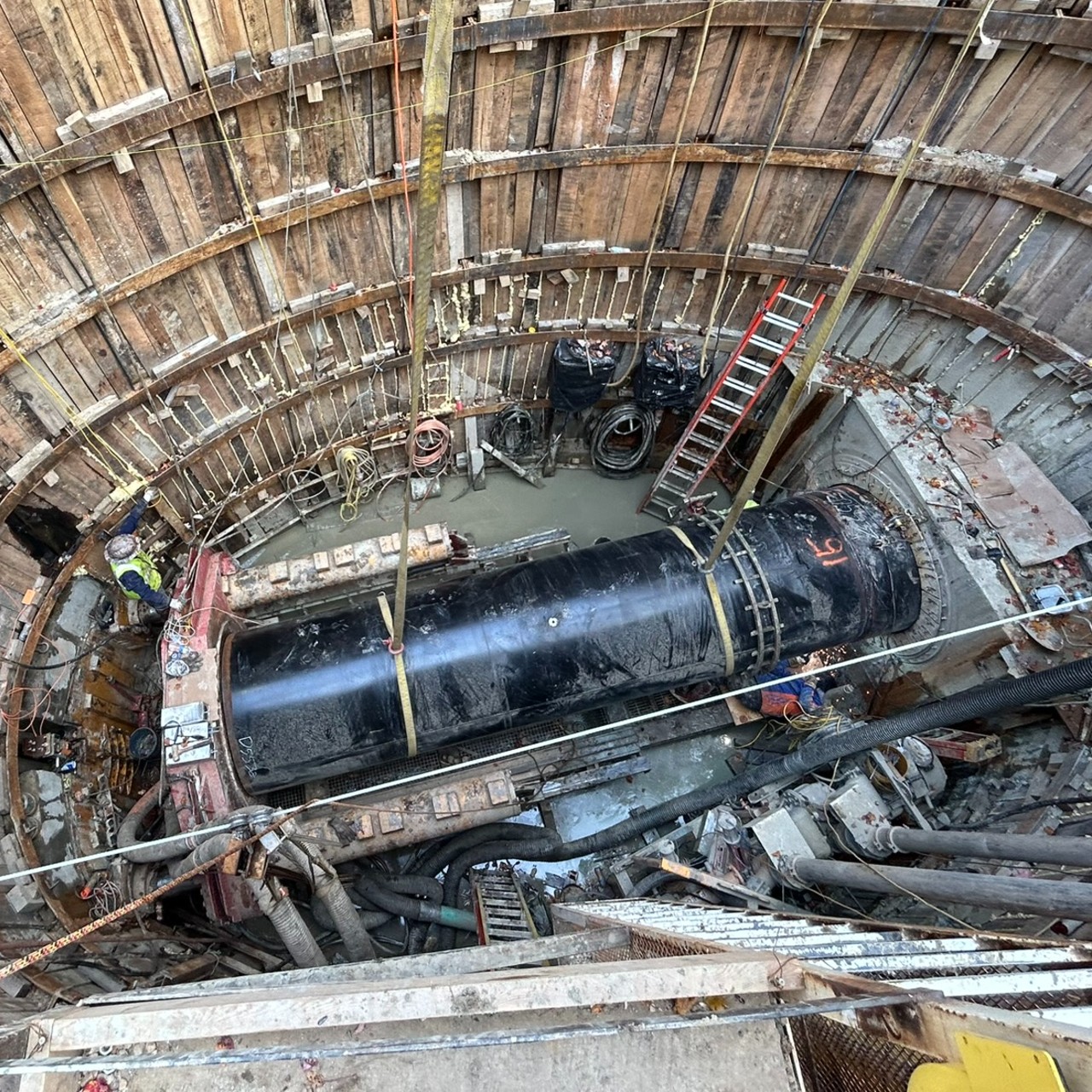 Looking down into a large hole at a construction site where there is a big black pipe supported by straps going into a tunnel.